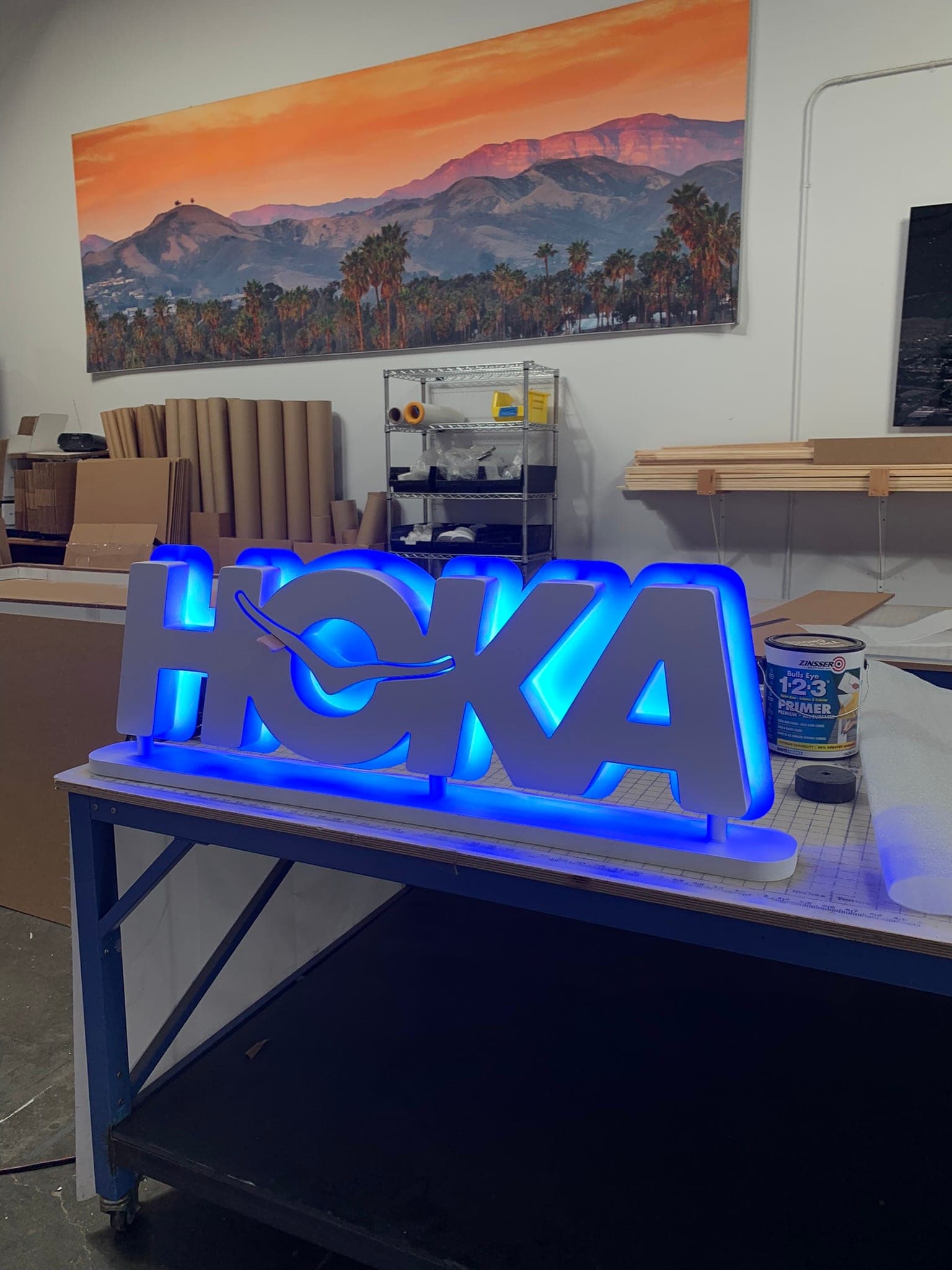 Backlit dimensional HOKA logo letters with a blue LED halo on the workshop bench, mountain landscape print on the wall behind.