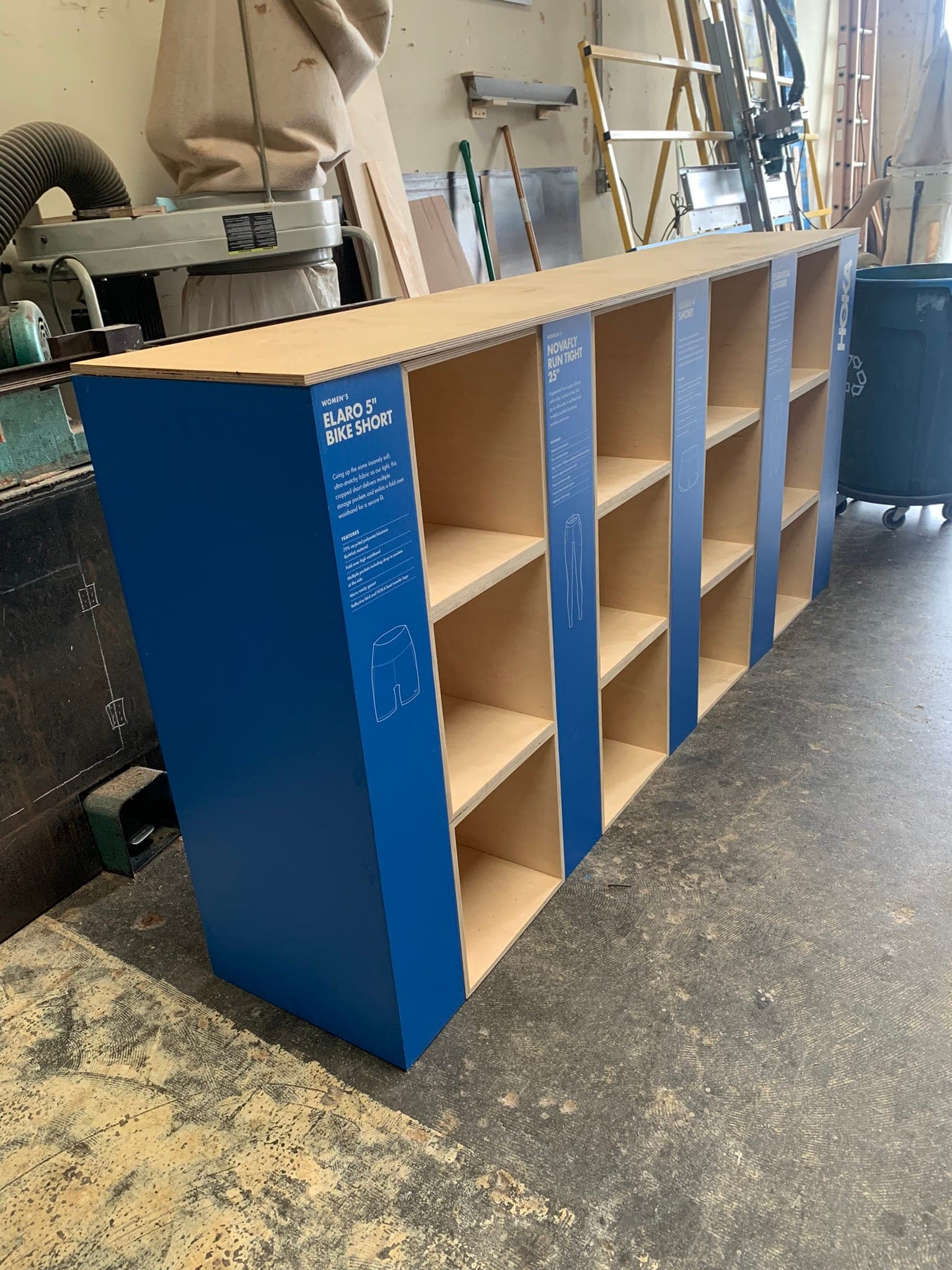 HOKA retail cubby fixtures — blue side panels with product spec graphics, raw plywood interiors, lined up on the shop floor.